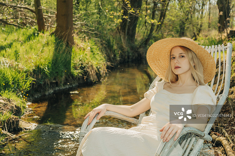 Woman relaxing in a wooden armchair by a country river wearing a straw hat图片素材