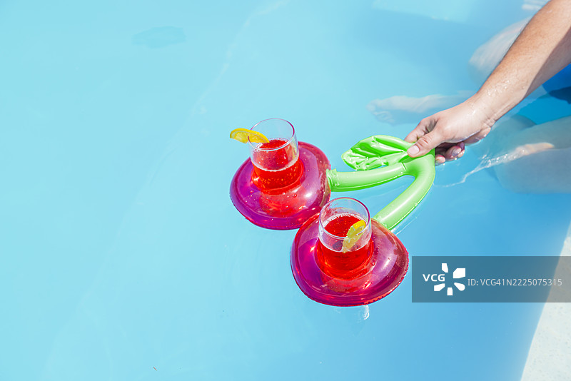 阳光明媚的泳池中，男人的手握着樱桃形的泳池浮具，旁边放着饮料。图片素材