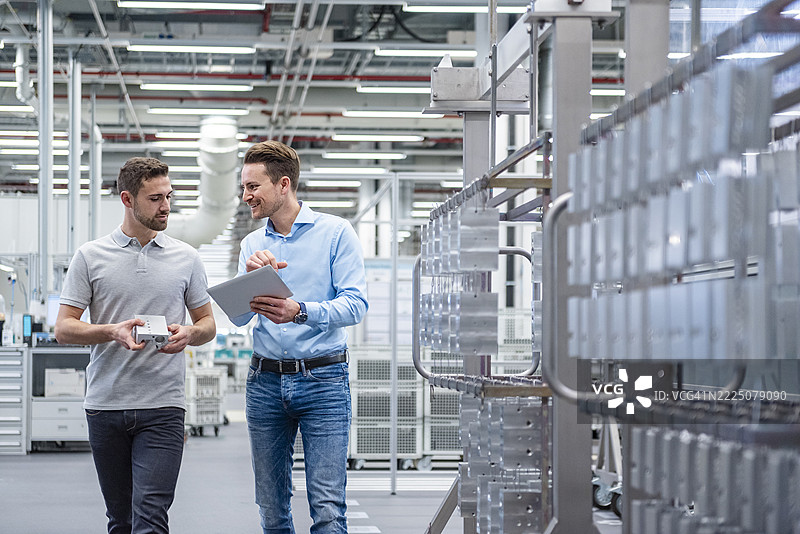两名员工在现代化的生产大厅中讨论，周围有机器和一台平板电脑。图片素材