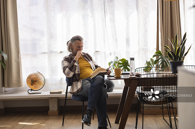 在家办公环境中，男人在自然光和植物的陪伴下阅读一本书图片素材