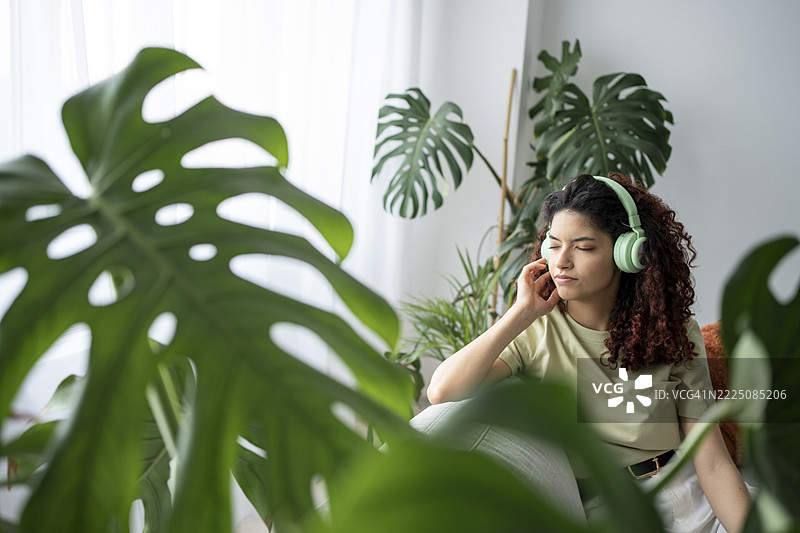 戴着耳机的卷发女性在室内被龟背竹环绕，沉浸在音乐中。图片素材