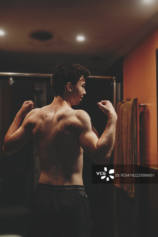 自信的年轻男性在镜子前赤裸上身，弯曲手臂展示肌肉。这个青少年面露专注神情，观察自己的倒影，象征着自我形象、个人成长或健身目标。图片素材