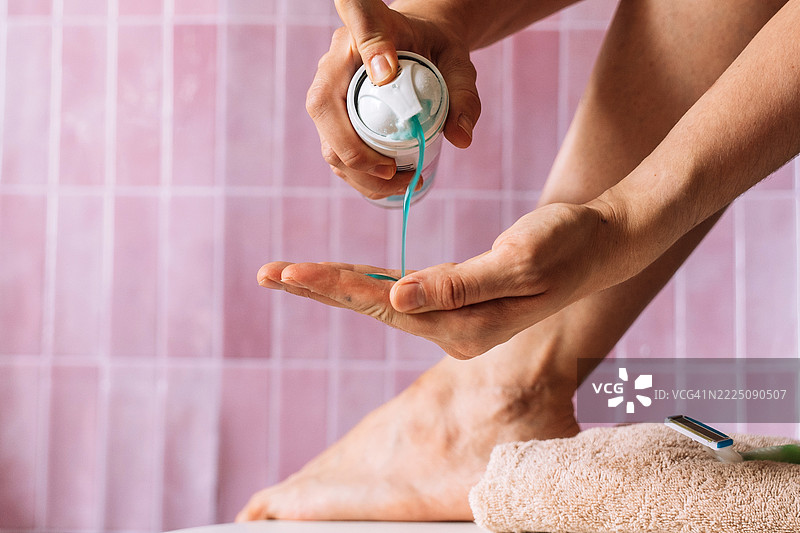 女性将蓝色剃须胶挤入手掌特写，背景为粉色瓷砖和双脚。图片素材