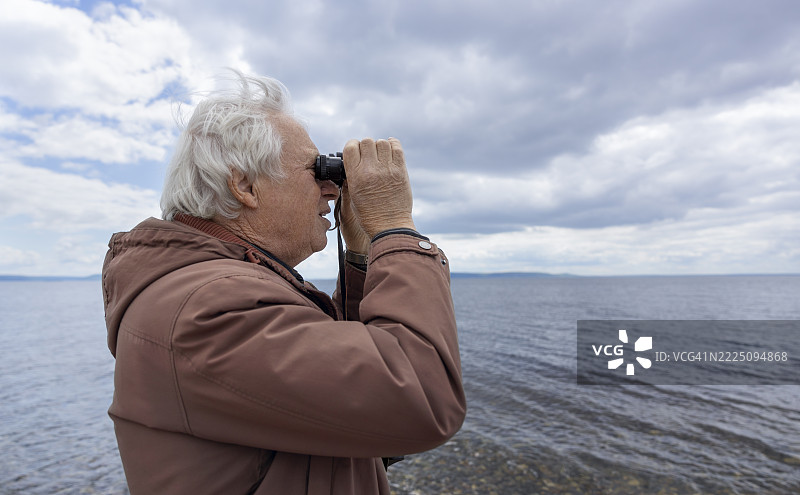 一位老年男子在雷雨交加的天空下，透过望远镜观察海滩。图片素材