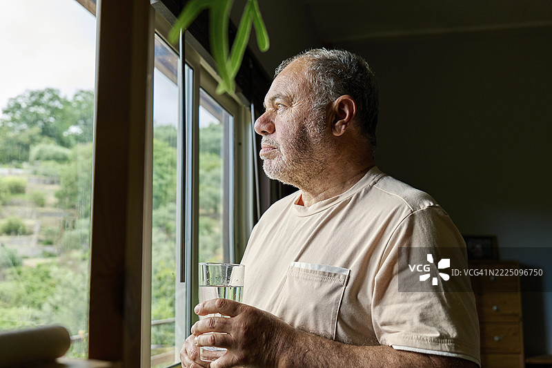 老年男子在家中望向窗外，手中拿着一杯水。图片素材