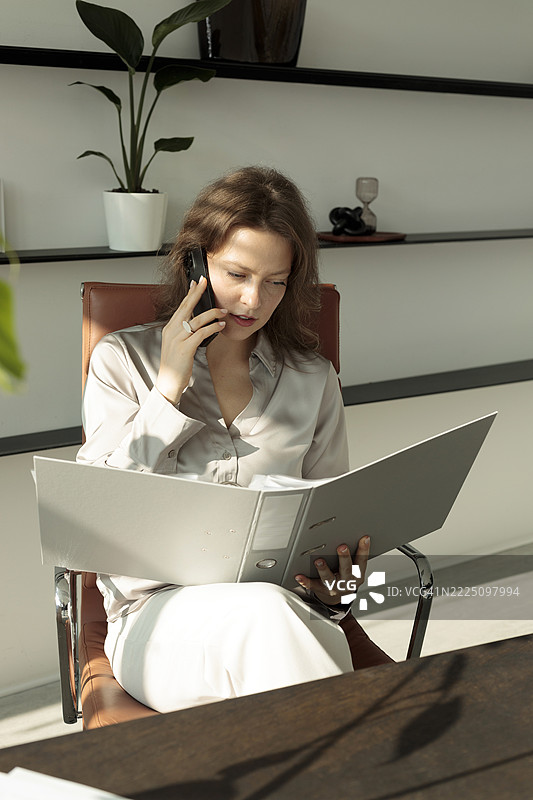 在现代办公室里，女商人一边打电话一边阅读文件图片素材