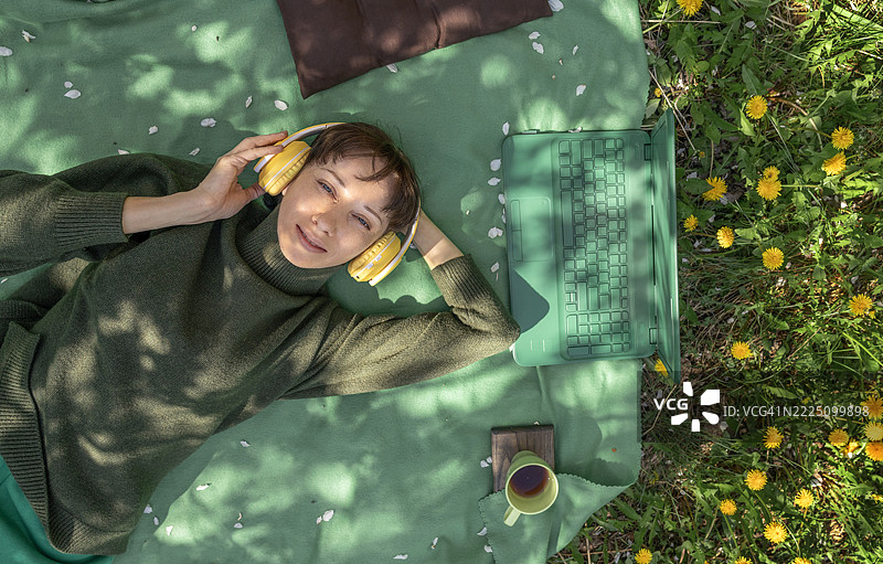 躺在草地上，戴着黄色耳机的女人一边听音乐一边使用笔记本电脑图片素材