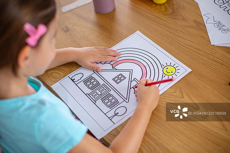 一个小女孩用彩色铅笔为一幅画着房子、彩虹和太阳的图画上色。图片素材