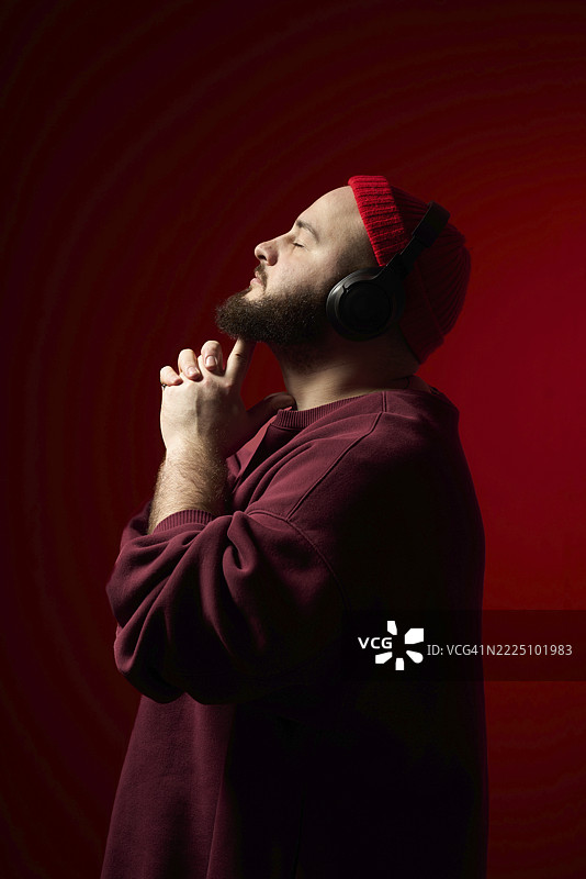 年轻男子佩戴耳机和红色毛线帽，背景为红色的工作室肖像图片素材