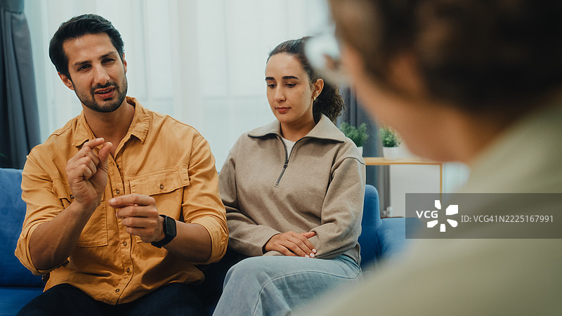 年轻夫妇在家中的客厅沙发上与心理健康专业人士进行严肃讨论，参加婚姻咨询会议。治疗会议心理健康概念。图片素材