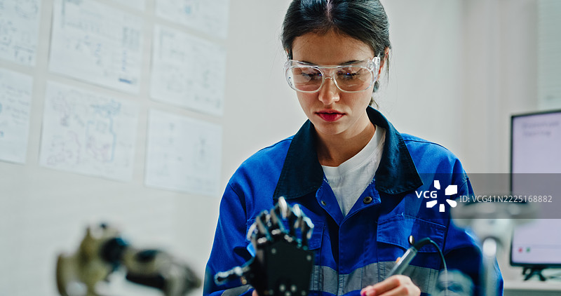 年轻女性工程师在技术实验室使用万用表和焊接工具测试和修理机器人手臂。人工智能与机器人开发。图片素材