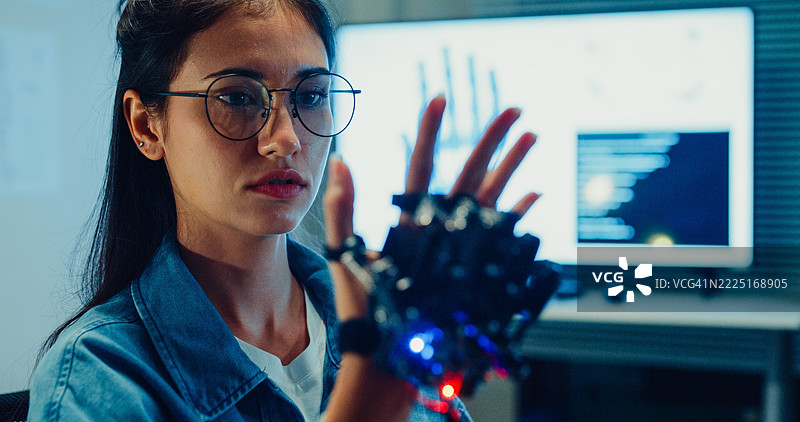 年轻女性工程师在夜间的科技实验室加班，向机器人手臂做手势。机器学习、计算机制造、创新与创造力。人工智能与机器人开发。图片素材