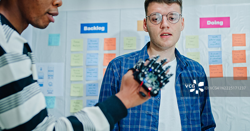 两名男性软件工程师在办公室仔细检查一个机器人手原型，进行开发和编码任务的集成。人工智能与编程。图片素材