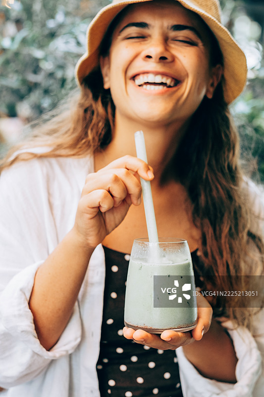 年轻女性在户外自然光下微笑着搅拌一杯绿色果昔，穿着波点裙和白衬衫。图片素材