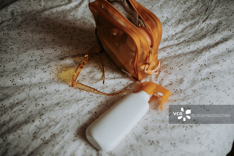 一个橙色的洗漱包放在床上，打开着，里面是夏季美容必需品。旁边是太阳镜和一瓶防晒霜。图片素材