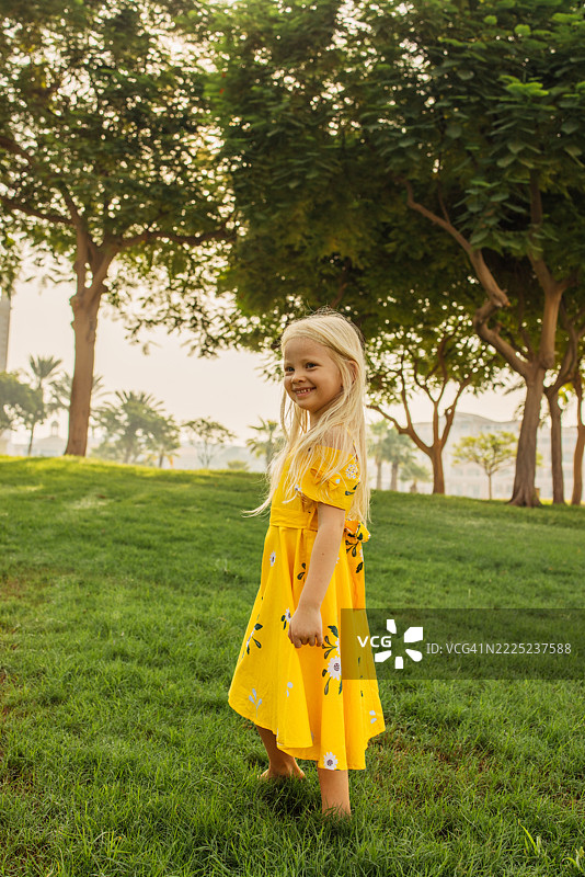 穿着黄色裙子的微笑小女孩站在阳光明媚的绿色公园里图片素材