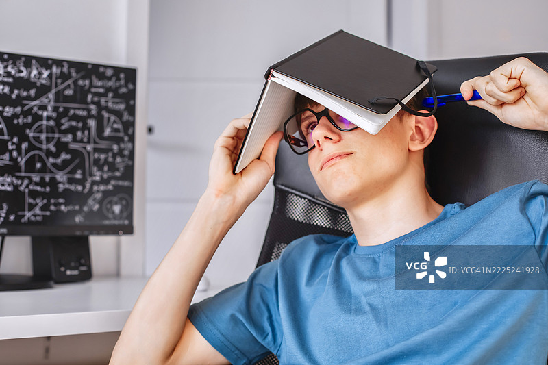 一个戴眼镜的青少年学生坐在电脑前，头上盖着一本笔记本。图片素材