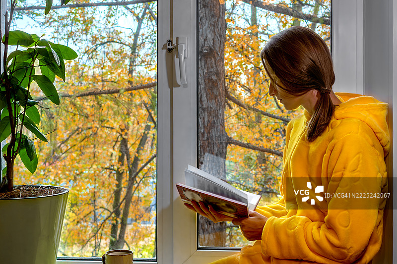 温馨的场景，一位穿着黄色浴袍的女性在窗边阅读书籍，窗外透出明亮的光线。图片素材