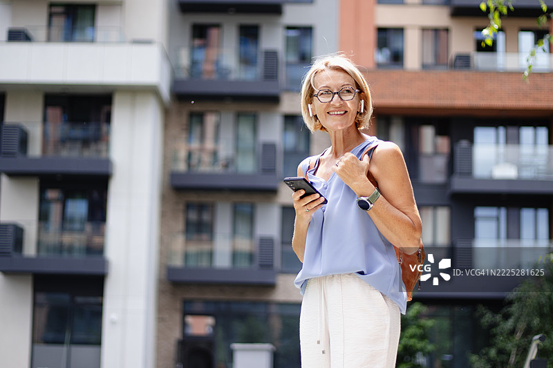 成熟的金发快乐女性戴着眼镜，在城市中散步，享受无线耳机听音乐，同时使用手机。图片素材