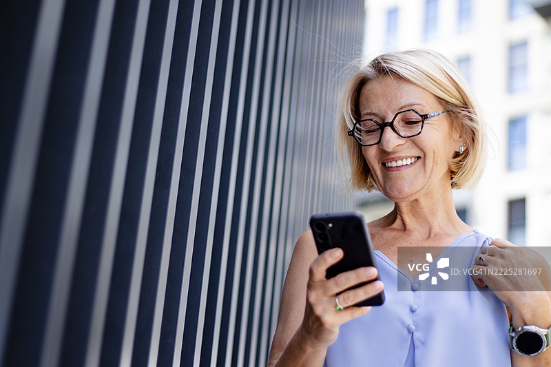 成熟的金发快乐女性戴着眼镜，在城市中散步，享受无线耳机听音乐，同时使用手机。图片素材