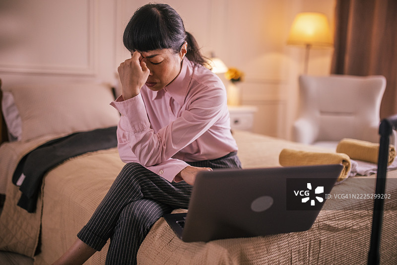在酒店房间里，压力重重的亚洲女商界高管正在分析笔记本电脑上的数据，并且在打电话。图片素材