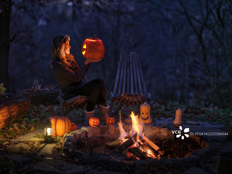 微笑的女性在万圣节晚上篝火旁与雕刻的南瓜。图片素材