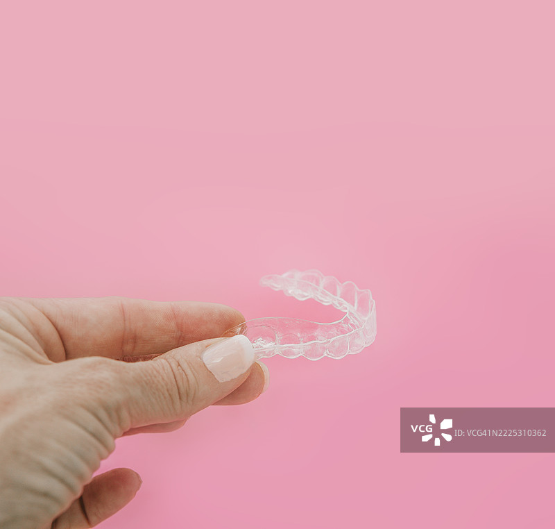一只女性的手在柔和的粉色背景下握着透明的正畸矫正器或保持器的特写。干净现代的构图非常适合牙科诊所的广告。图片素材