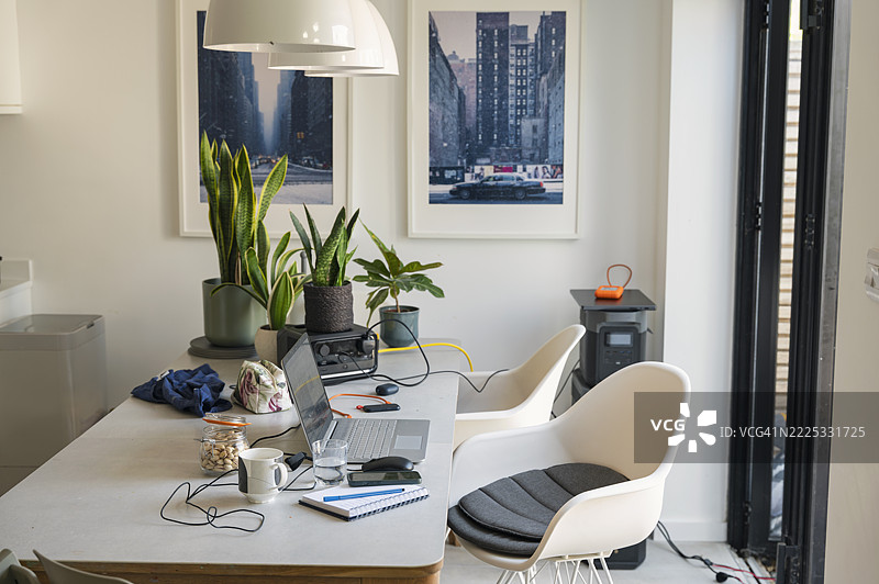 餐桌上的静物摄影，展示了一台笔记本电脑，处于居家办公环境中。它由一个与太阳能电池板（镜头外）相连的电池储存系统供电 - 可持续能源。图片素材