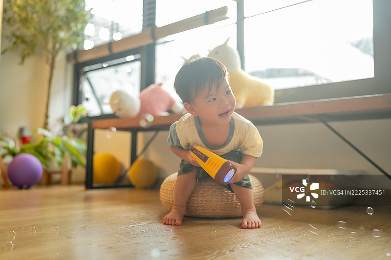 亚洲小男孩在客厅里快乐地玩耍，抓住肥皂泡，周围弥漫着清新愉快的夏日气息。孩子开心地笑着，泡泡在空中飘荡，营造出一个有趣而活泼的室内时刻。图片素材