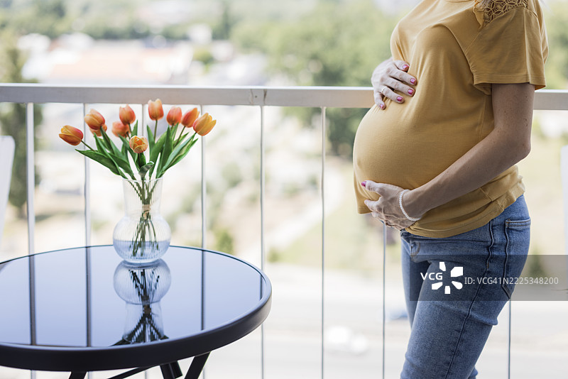 一位孕妇站在阳台上，手抚摸着腹部，特写镜头图片素材