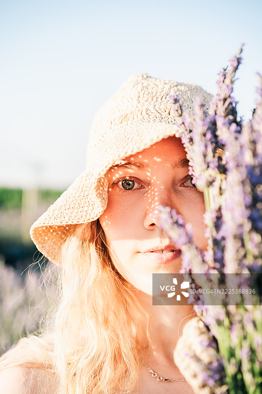 戴着草帽的女人柔和肖像，手捧薰衣草，柔和的阳光与自然之美。图片素材