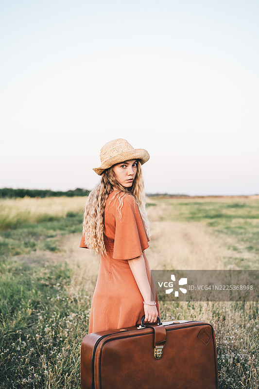 穿着橙色连衣裙的女性复古旅行肖像，手持行李箱，乡村小路和柔和的日落光线。图片素材