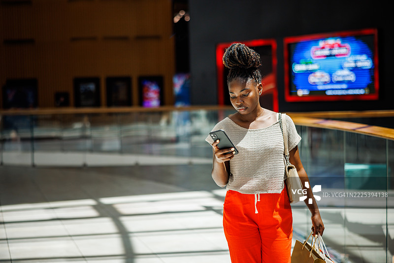 年轻女士在商场里一边走一边使用智能手机，手里提着购物袋图片素材