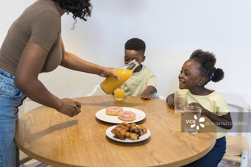 妈妈在早餐时为她的两个孩子倒橙汁，桌上放着甜甜圈和糕点图片素材