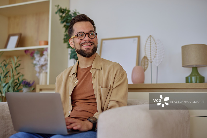 年轻男子在家中沙发上工作，使用笔记本电脑，微笑着望向一旁。图片素材