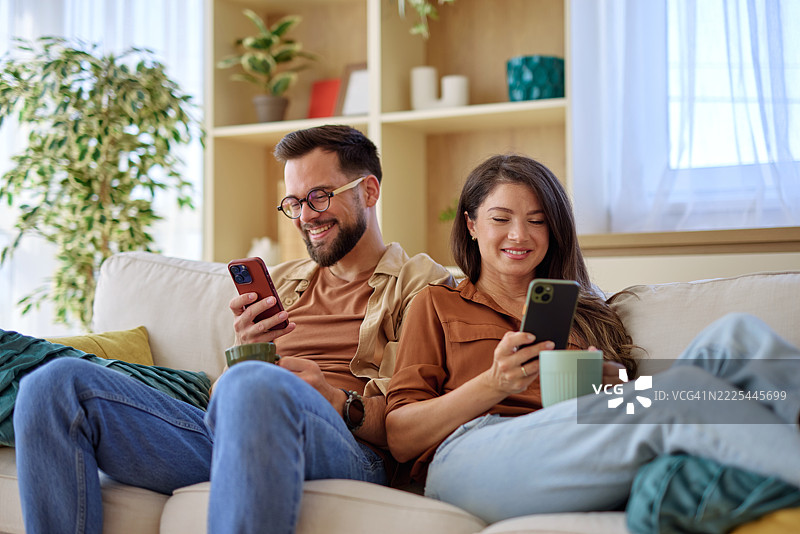 年轻情侣在沙发上放松，浏览智能手机并喝咖啡图片素材