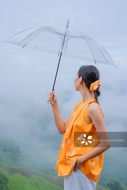 年轻女性穿着明亮的橙色上衣，手持透明雨伞，站在雾气弥漫的绿色山丘风景中，雨天的场景。图片素材