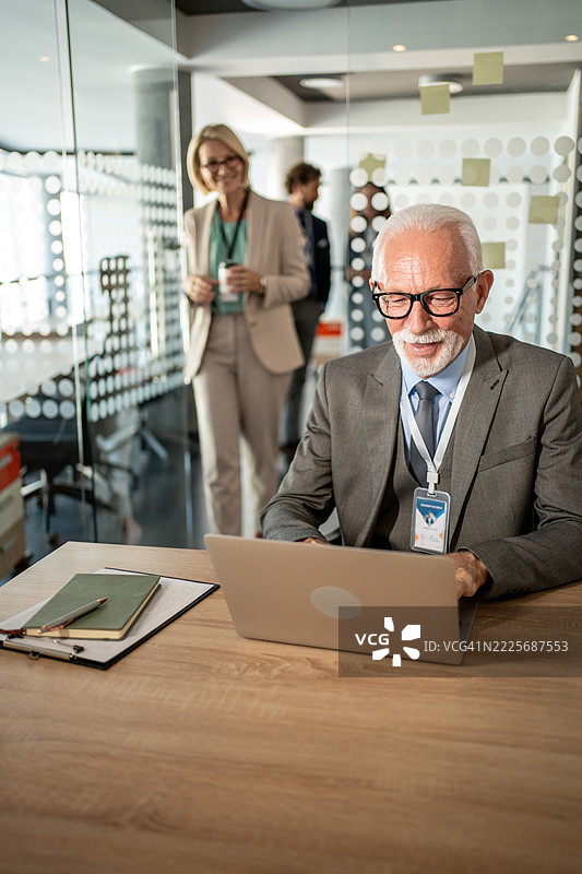 一位高级商务人士在办公室使用笔记本电脑，背景中同事们正在交谈。图片素材