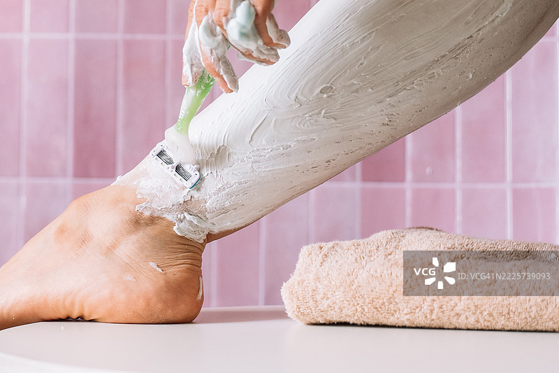 女性腿部涂抹剃须膏，泡沫覆盖，剃刀从脚踝开始，背景为粉色瓷砖。图片素材