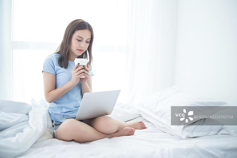年轻女性坐在床上使用笔记本电脑并拿着杯子图片素材