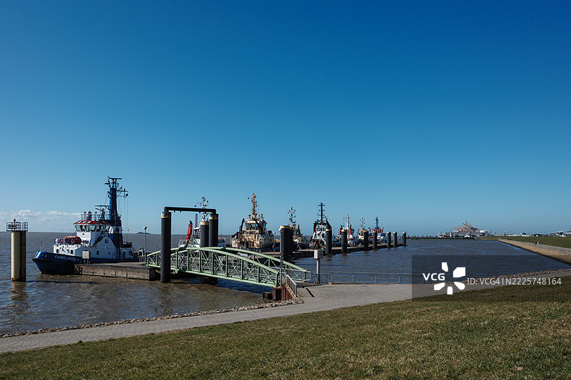 一幅宁静的画面，展示了一队各种拖船整齐地并排停靠在平静港口的长码头上，蓝天晴朗，阳光明媚。图片素材