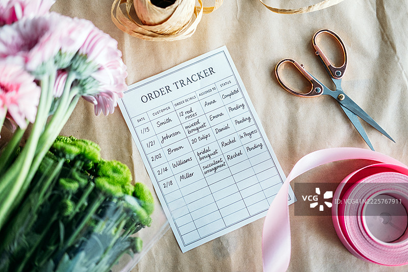 花店订单跟踪表的特写，周围有丝带、剪刀和新鲜花朵，铺在牛皮纸包装上。花卉业务规划、订单管理、花店工具、手工花卉服务图片素材