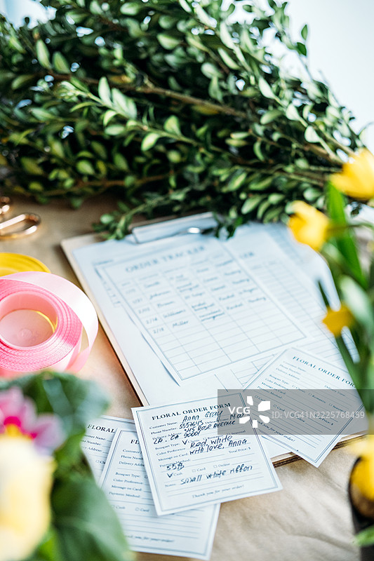 花店工作区内，手写的花卉订单表格特写，配有夹板、丝带和绿植。客户体验个性化、手写订单、定制花艺、精品花卉业务。图片素材