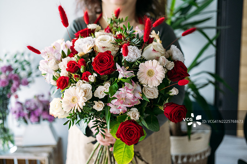 女人手持优雅的红白玫瑰、康乃馨和绿植花束，背景为白色。适用于婚礼和周年庆典的活动造型，经典花卉设计，新娘花束，浪漫庆祝活动。图片素材