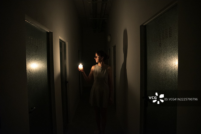 年轻女子在停电时手持点燃的蜡烛走过黑暗的走廊。图片素材