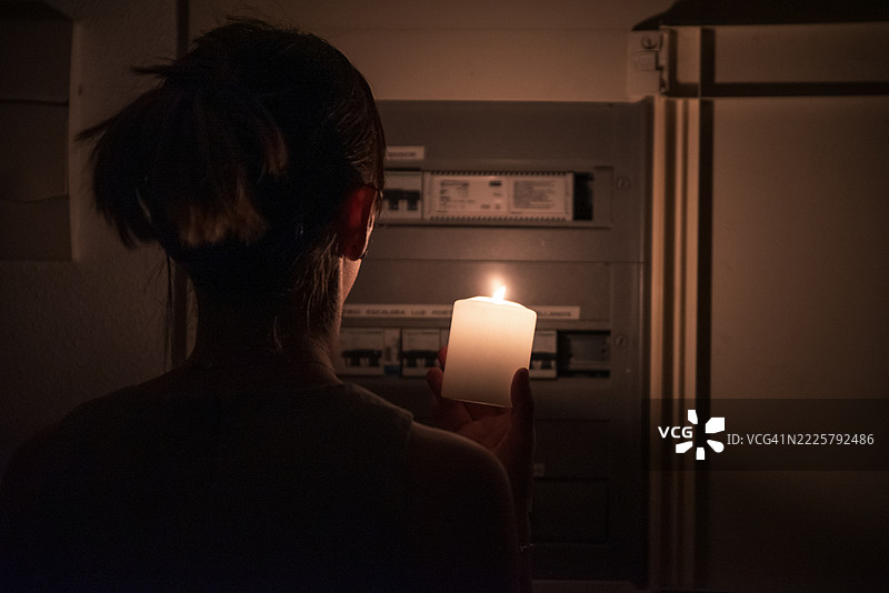 一位面容模糊的女性手持点燃的蜡烛，检查电气面板是否有电。图片素材