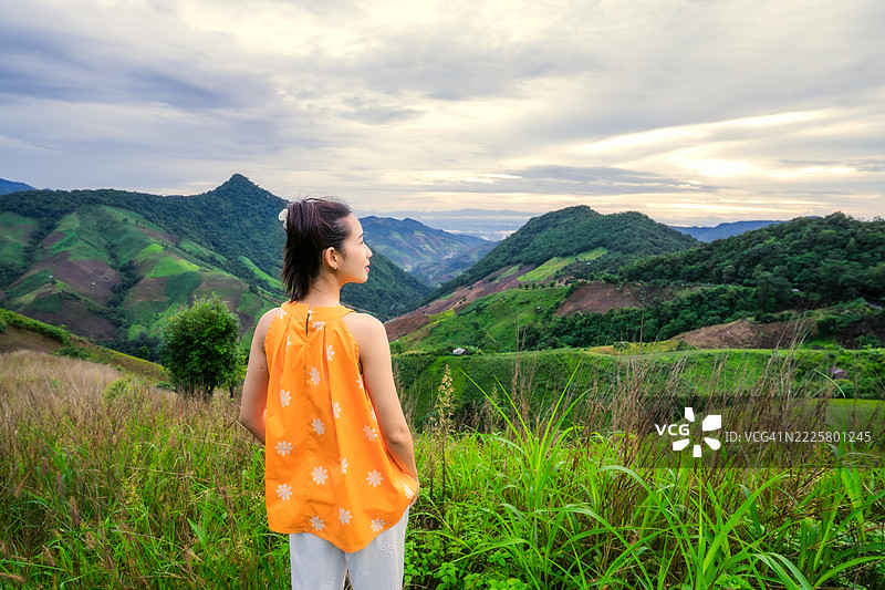 年轻女性站在高草中，欣赏着壮丽的绿色山脉景观，背景是戏剧性的多云傍晚天空。图片素材