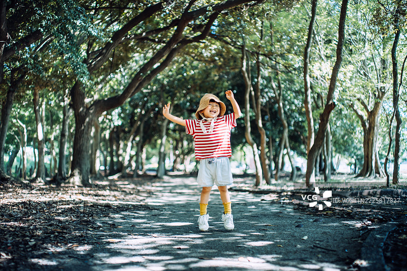 一位快乐的小亚洲女孩戴着草帽，在公园的森林小径上欢快地跳跃。享受大自然的乐趣，孩子们在户外玩得开心。图片素材