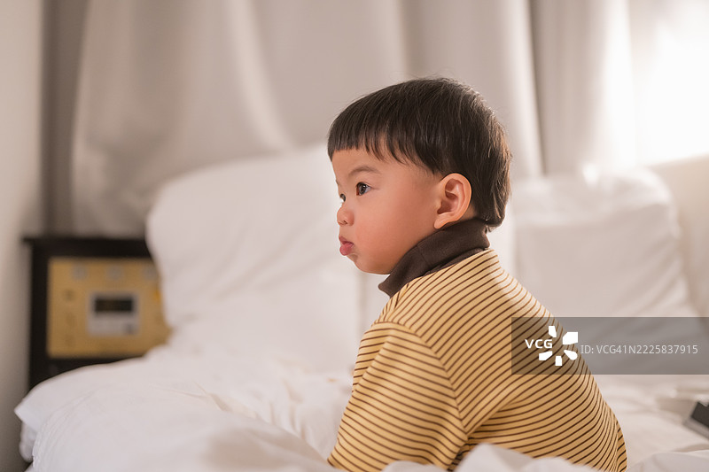 好奇可爱的亚洲幼儿男孩坐在酒店房间的床上，正在爬行，身边有一个树盆玩具。图片素材