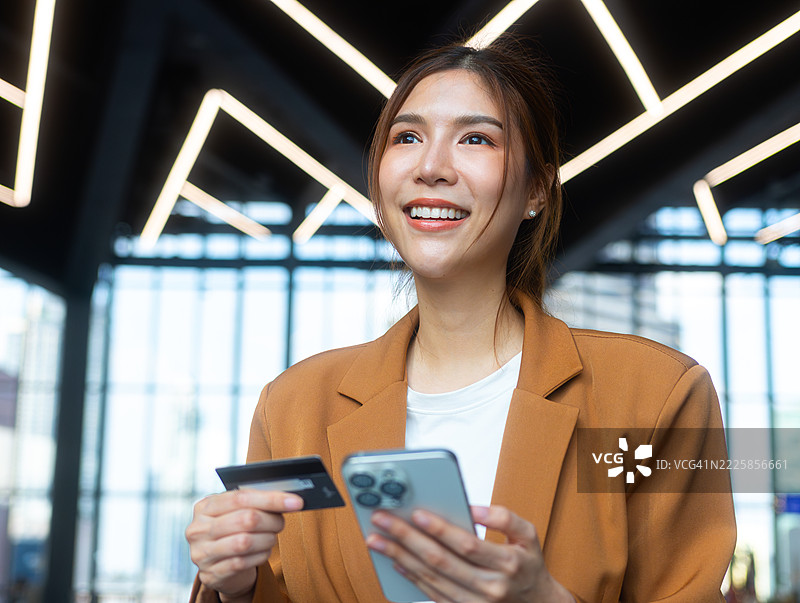 自信的亚洲女商人手持信用卡和智能手机，微笑着在现代办公大楼中使用移动银行应用进行在线支付和数字金融服务。图片素材